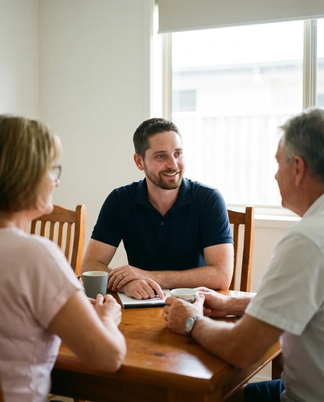 Vertical photograph of Middle Park Financial Adviser Jonathon De Martini smiling and listening to clients at a sunlit wooden dining table.
