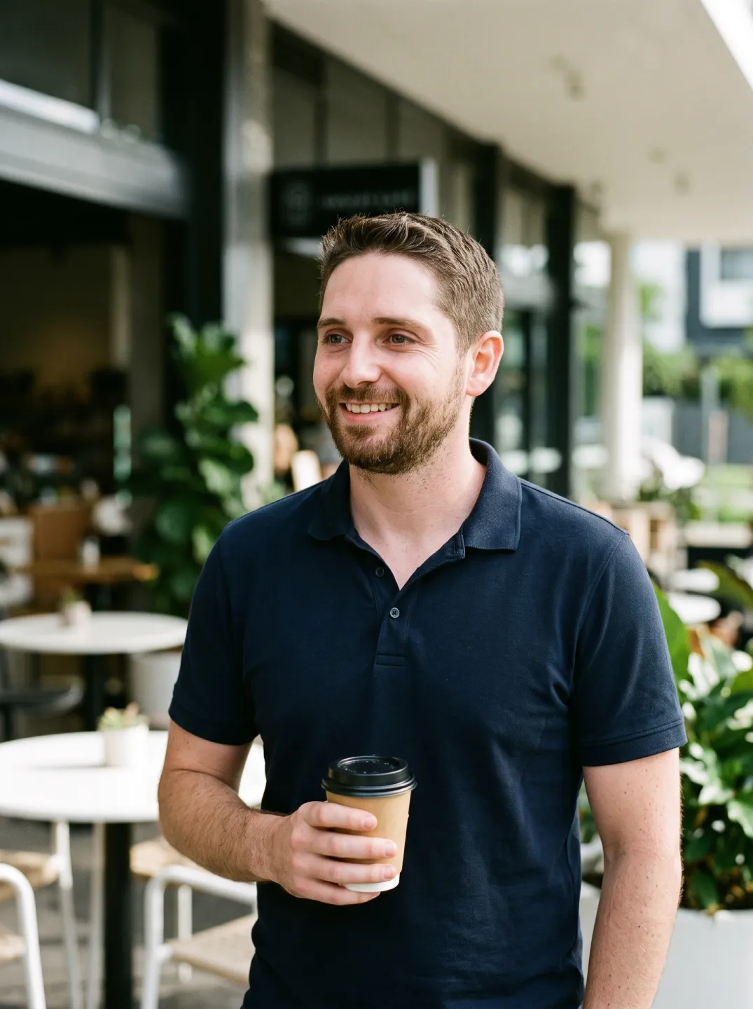 A professional photograph of Jonathon De Martini, a Financial Adviser in Middle Park, Brisbane, smiling and holding a takeaway coffee outside a leafy modern cafe.