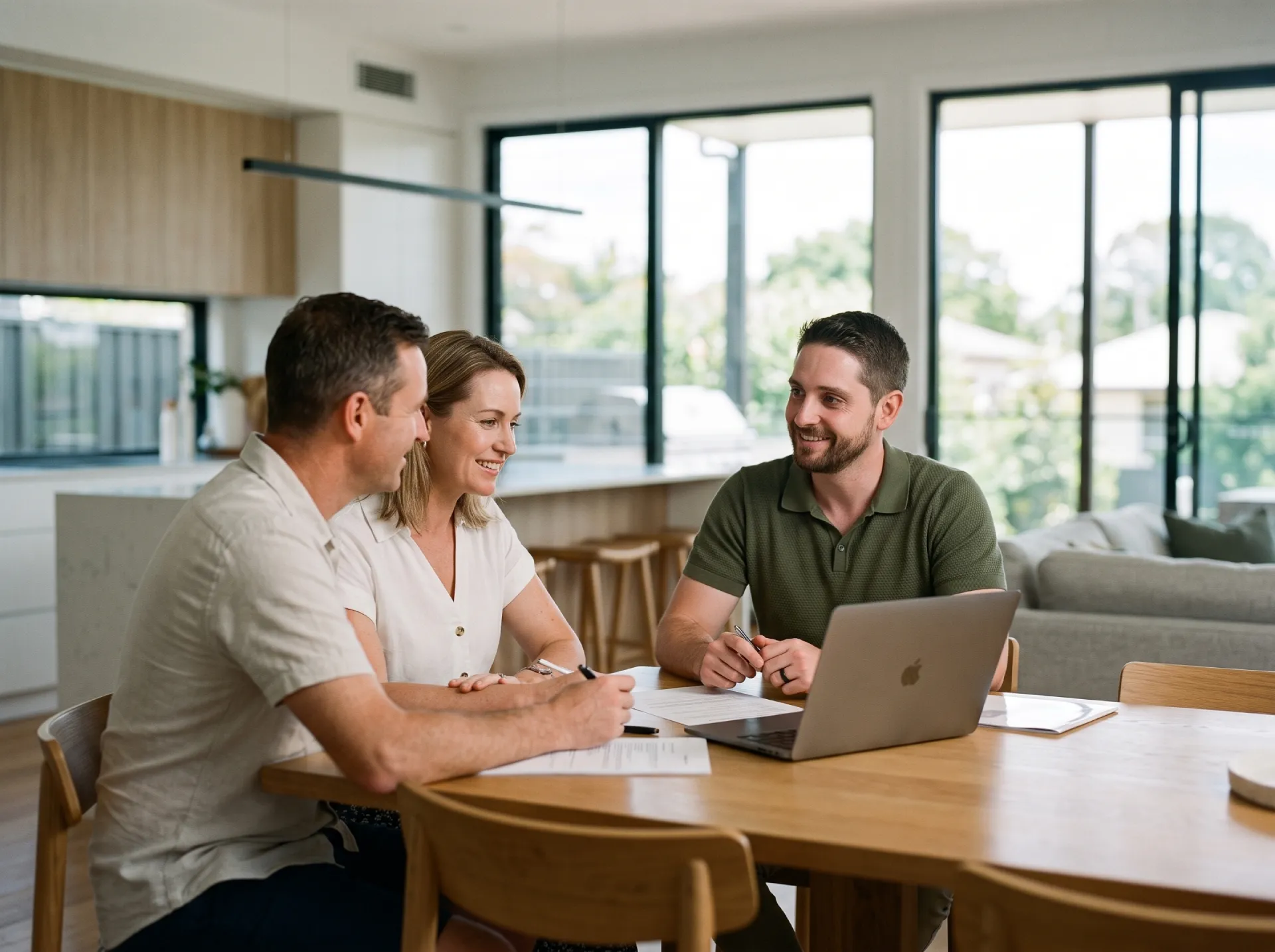 Jonathon De Martini, a Financial Planner from Middle Park Brisbane, smiling and presenting financial information on a laptop to a couple in a light-filled Brisbane home setting. A wooden table with a laptop, notepad, and documents is visible.