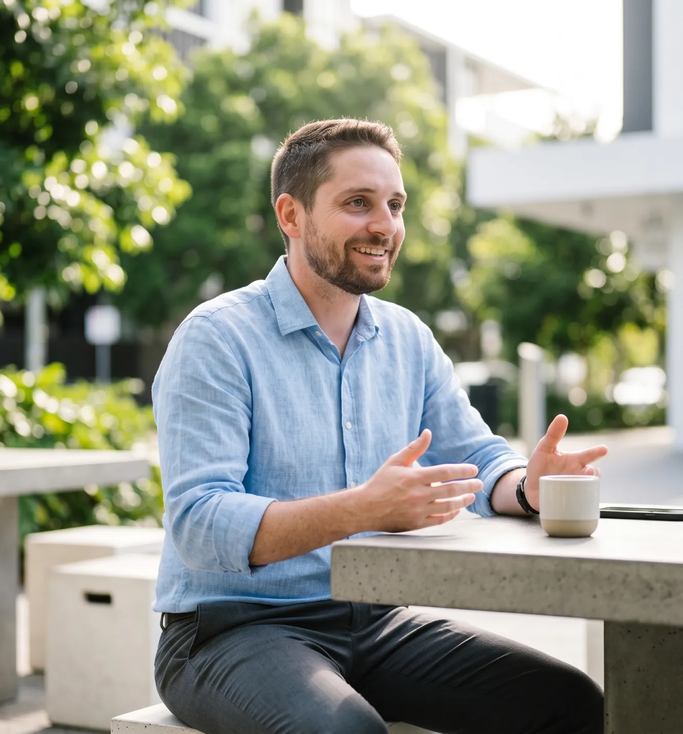A professional photograph of Jonathon De Martini, an authorised financial planner based in Middle Park Brisbane, conducting a superannuation advice consultation with a diverse couple. He is smiling and gesturing towards an iPad Pro screen showing modern financial data visualisations, set at an oak table in a modern office. The background features a large window view of the Brisbane River and city skyline under natural golden hour light.
