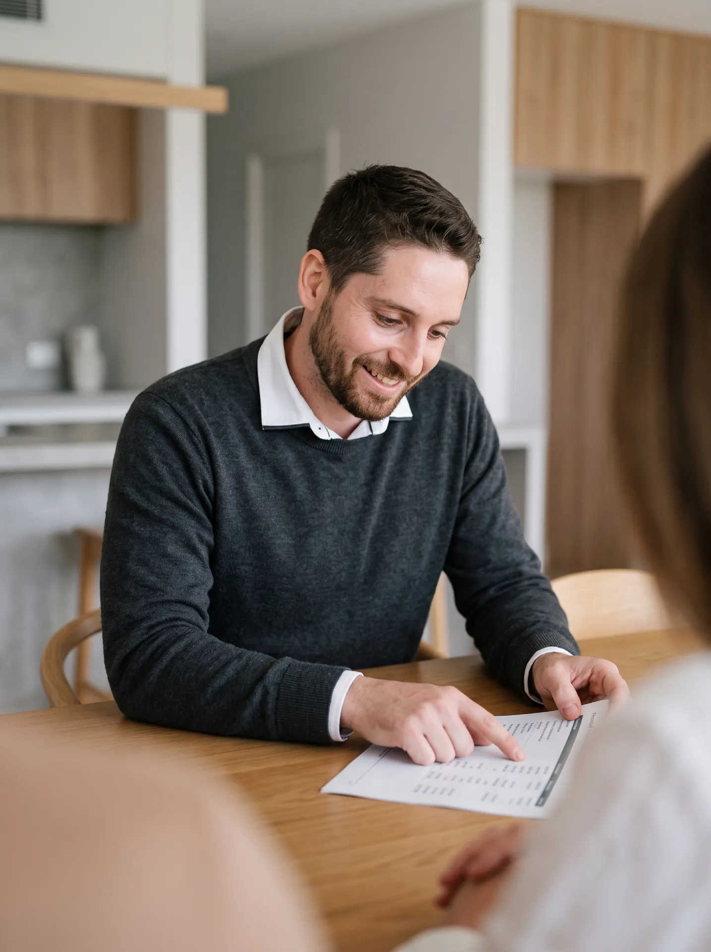 Jonathon De Martini, a Brisbane Financial Planner in Middle Park, points to a financial document while thoughtfully explaining wealth accumulation strategies to a client at a wooden table in a bright, modern Brisbane home.